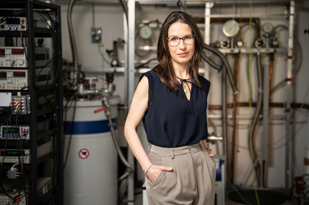 Image of Czech physicist Helena Reichlova, standing in a lab, wearing a black short-sleeved blouse and grey chinos, in front of technical equipment.