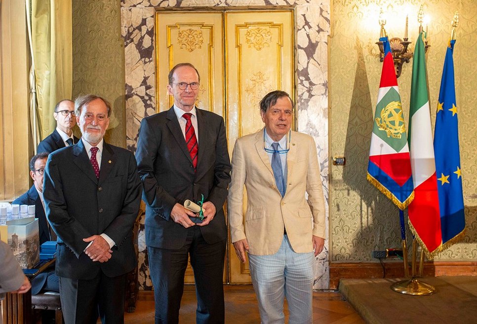 Three gentlemen in suits, on the right three flags, including those of Italy and Europe